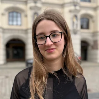 Luna Spiegel blickt direkt in die Kamera, mit dunkler Brille und schwarzem Oberteil, im Hintergrund die helle, verzierte Fassade eines Gebäudes.